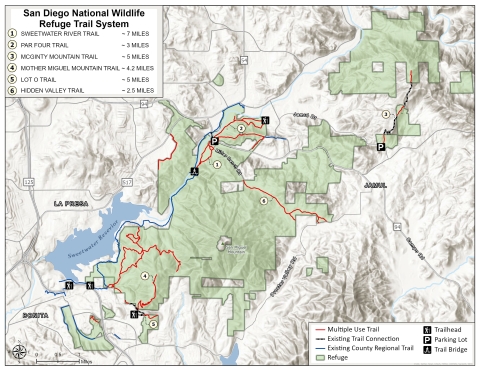 The map shows the trail system for San Diego NWR. On the upper left is a box with the names of six different trails. The names are numbered, and the number matches where on the map the trail is located. The refuge boundaries, in green on the map, show multiple use trails, trail connections, county trails, and icons show where the trailheads are, parking lots, and trail bridge. For more map information, contact the refuge manager 619-385-1190.