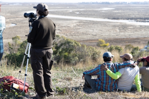 Four people are pictured from behind, looking out over a winding waterway. One person is taking photos and another is looking through binoculars.