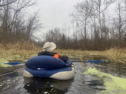 On a chilly day, a person floats down a swampy waterway surrounded by dried foliage and leafless trees.