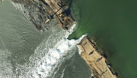 An overhead shot of a bulldozer removing a portion of an earthen levy between two bodies of water. 