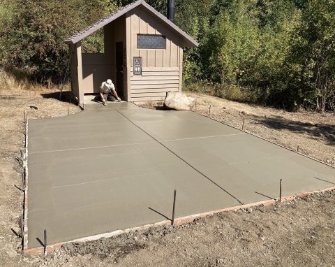 A new concrete pad is poured near a vault toilet as a worker smooths its surface with a hand tool.