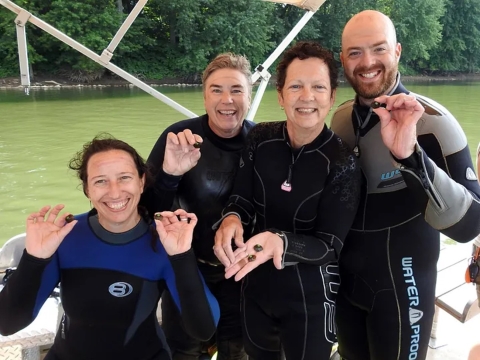 Group photo of four biologists on a boat, wearing diving suits and holding freshwater mussels that they are about to release into the water.