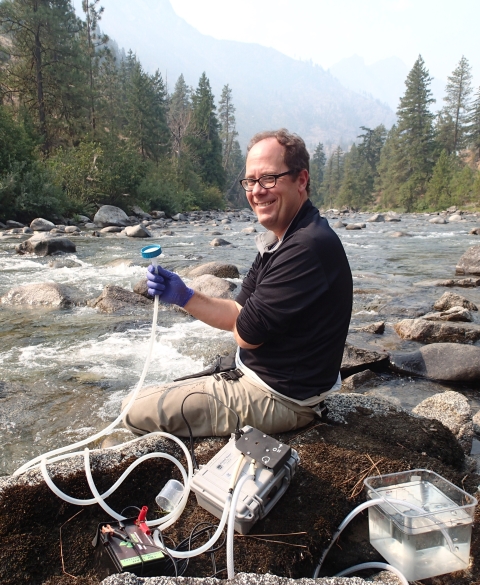 A man wearing glasses sits on rocks at a river's edge, smiling and holding a plastic tube connected to field equipment. Trees and a mountain are in the distance.