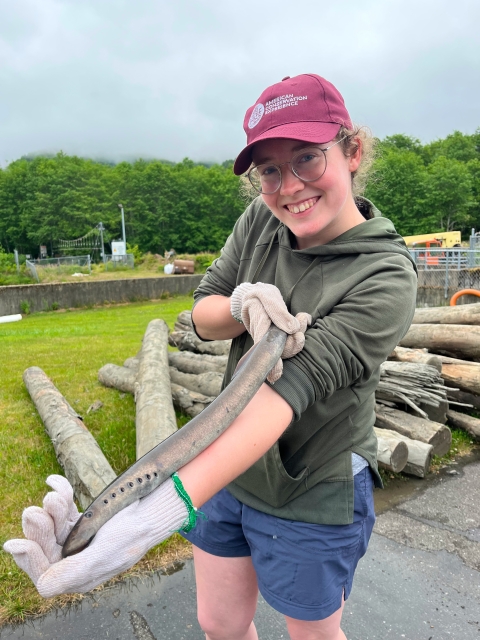 A woman smiles while holding a Pacific lamprey. Logs and structures from Makah National Fish Hatchery are in the background below the trees and sky.