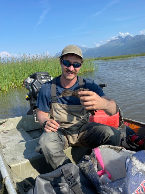 A smiling person wearing chest waders, sunglasses, and a brimmed hat sits in a boat holding a duck