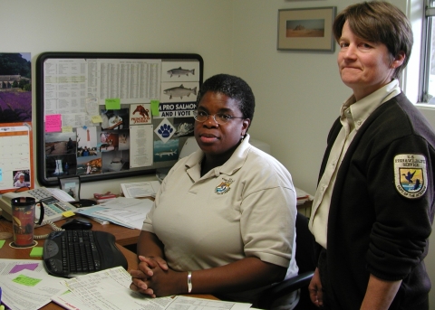 Two women in an office looking at the camera. One is sitting behind a desk with her hands folded and one is standing next to her.