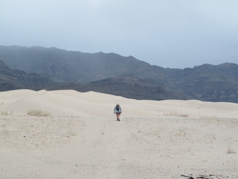 a woman walks across white sand dunes headed toward rugged mountains