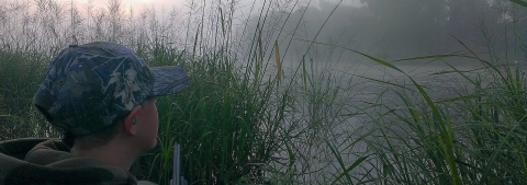 A young hunter in a camouflage cap crouching in talk grass next to a body of water