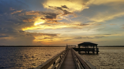 the sun shinning through the clouds as it sets over the water. A long wooden dock to the boat lift in the foreground.