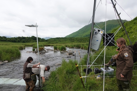 Three people in USFWS uniform. Two are erecting a light over a river while the third observes.