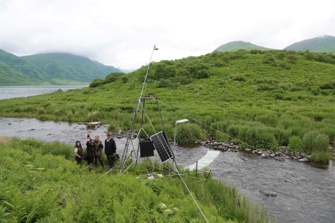 Four people in USFWS uniform wave or smile to the camera from next to a fish monitoring station next to a river.