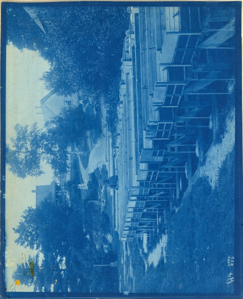 A historic 1870's photo shows a line of wooden troughs with a man standing at the rear in bibs and a hat. Behind the man is a hill with trees and two houses on it, and two women wearing white dresses walking up the hill.