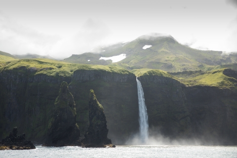 Waterfall plunges into the Bering Sea over a high cliff with foggy, snowy, and green mountain in the background.