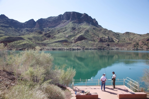 Two people fish at a pier with mountains in the background.