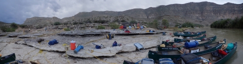 A campsite containing camping and scientific monitoring equipment is shown next to a river containing several canoes.