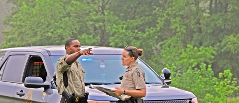 Two law enforcement officers -- a man and a woman -- speaking as the male officer points. They are in front of a police car in a wooded setting.