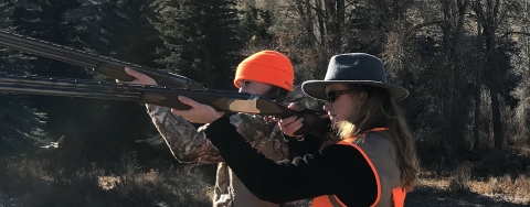 Two women in hunting gear side by side holding long guns side in shooting position