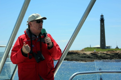 Man stands at the front of a ship holding a microphone. A lihthouse is seen behind 