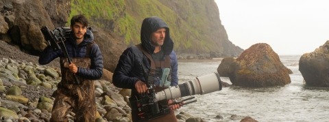 Two men carrying videography equipment walking on a rocky beach