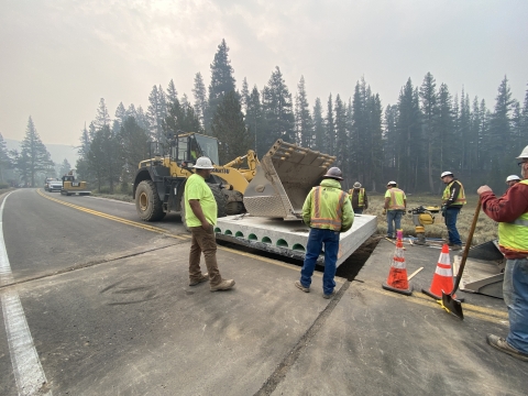 Construction workers install culvert in mountain road with a background of pine trees under a hazy sky.