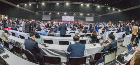 A fish-eye shot of an international convention, many tables, flags, and people having conversations, indoors.