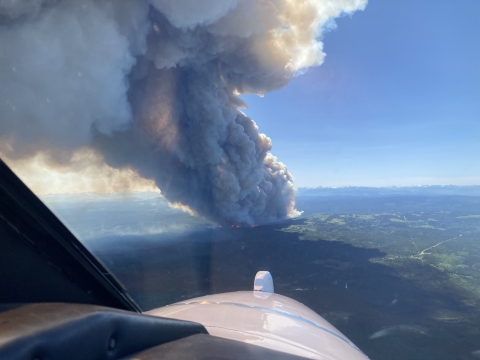 A view of a smoke plume from an airplane. The plume of smoke is billowing into blue sky from a forested landscape.