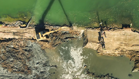 A bulldozer works to remove part of an earthen levy separating two bodies of water, while a group of people watch. 