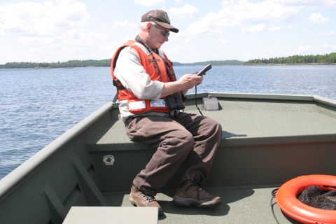 A man in an orange vest and refuge clothing sits on the bow of a boat in the middle of a lake looking at surveying equipment