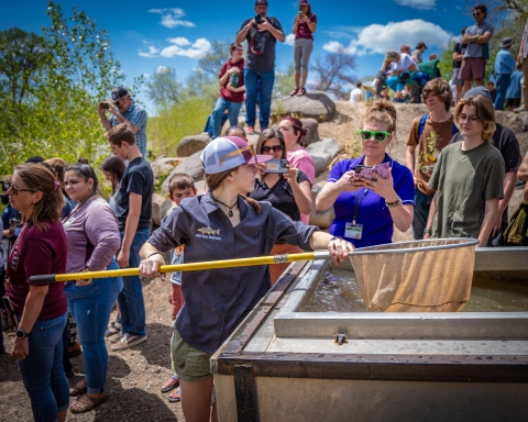 A high school student holding a net over a fish trailer. There are hundreds of people in the background as well as green trees and a blue sky with clouds.