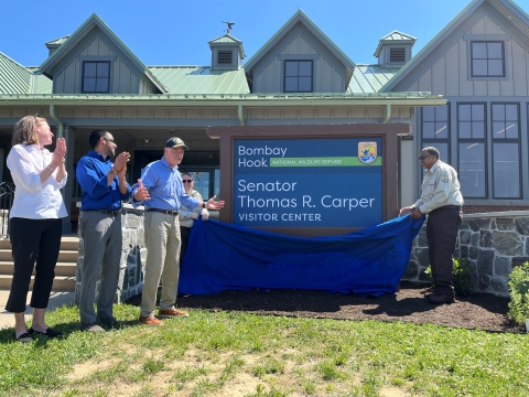 a group of people stand at a large visitor center facility. One of the people pulls off a blue fabric to unveil a sign that says "Bombay Hook Senator Thomas R. Carper Visitor Center". People are clapping in celebration of the event.