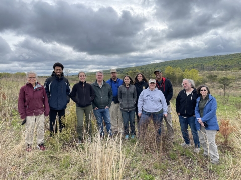 a group of 11 people stand amidst grasses in a landscape of rolling hills under gray skies