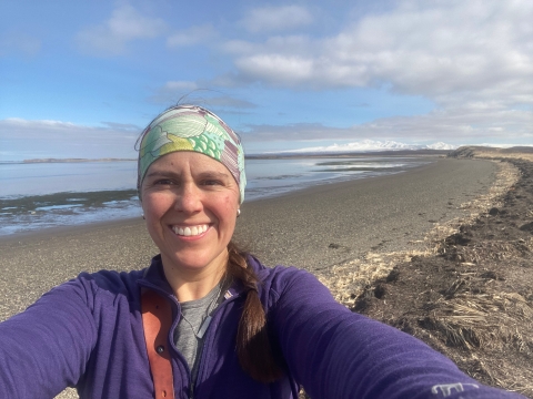 Maria Fosado smiles for a selfie on an ocean shore. Snow-covered mountains loom in the distance.