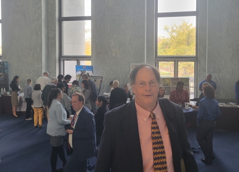 A white man with glasses and gray hair wearing a peach-colored shirt, patterned tie, and gray suit coat stands in a large room with people and windows in the background.