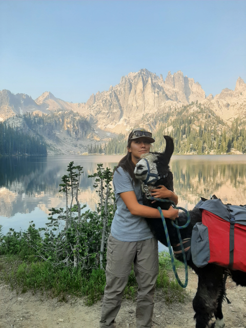 A woman wearing a ball cap hugs an alpaca carrying a pack in front of a still lake, blue sky, and big mountains.