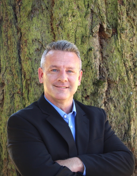 Person smiles for photo wearing dark suit jacket, standing in front of tree.