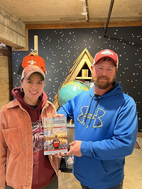 woman wearing a red hat, orange jacket and red hooded sweatshirt holding a book with a man wearing a blue hooded sweatshirt and red hat. World globe and bookshelf with dark wall in background