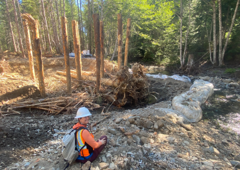 A woman wearing a white hard hat and bright orange safety vest smiles and sits along the side of a dry river restoration site with large wooden structures standing out of the riverbed.