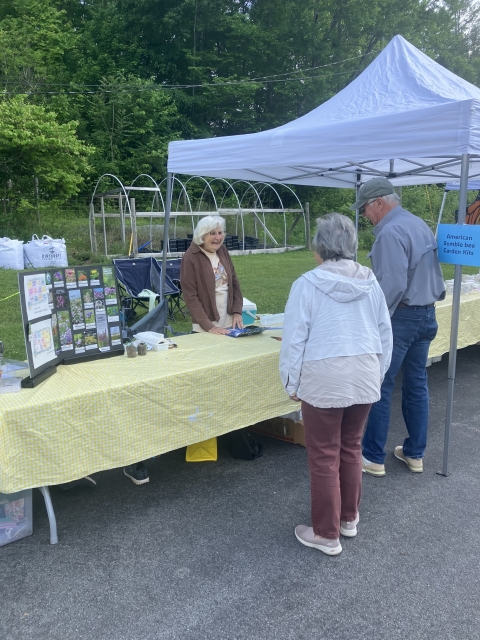 Two people are standing behind a table , talking to another lady who is beside a poster about native plants