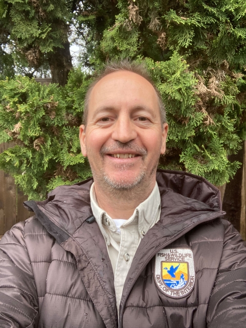 Headshot of a smiling man standing in front of green evergreen trees.