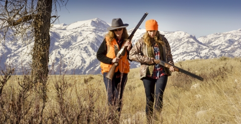 Two women carrying rifles and wearing bright orange gear walk through grass with the Teton Mountains behind them.