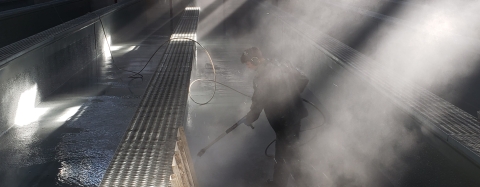 A photo of a person using a sprayer to clean tanks inside of a hatchery structure. There is a lot of mist in the air and light coming through the mist slightly obstructs the person. 