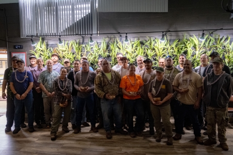 Members of the State of Hawaiʻi Department of Land and Natural Resources Department of Forestry and Wildlife fire management team gather in front of a plant wall at the Maui Bird Conservation Center. They are wearing lei and holding engraved bowls. 