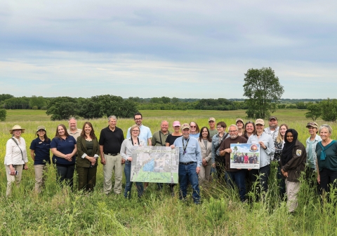 A group of people stand in a field facing the camera and holding up a map and another piece of printed material.