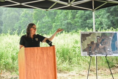 a woman wearing a black polo shirt stands behind a lectern and gestures toward a map, with grass and forest behind