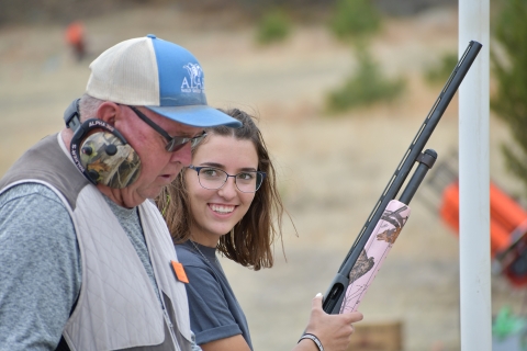 Youth participant in shooting sports holding pink firearm stands next to mentor. 