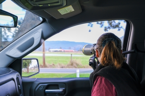 A biologist sits in the passenger side of a truck and peers through a spotting scope at birds in a field.