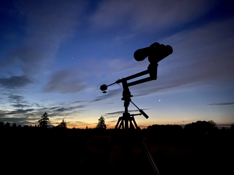 Astronomy binoculars in the foreground, with a sunset and dark sky in the background.