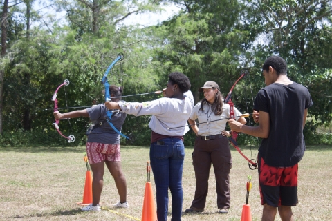 Three teenagers holding bows stand in a line receiving instructions from a uniformed FWS staff.