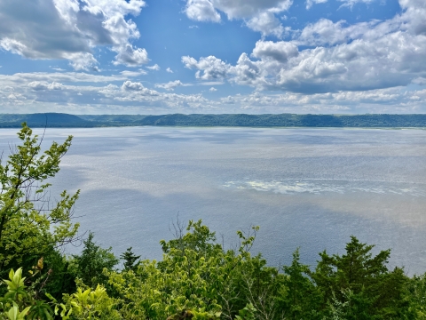 A blue sky with clouds as the sun shines on the upper Mississippi River with green bluffs in the distance.