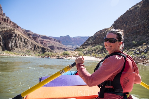 A woman wearing sunglasses and life jacket paddles in a kayak down a river in the Grand Canyon. She is looking back to smile for the camera. 